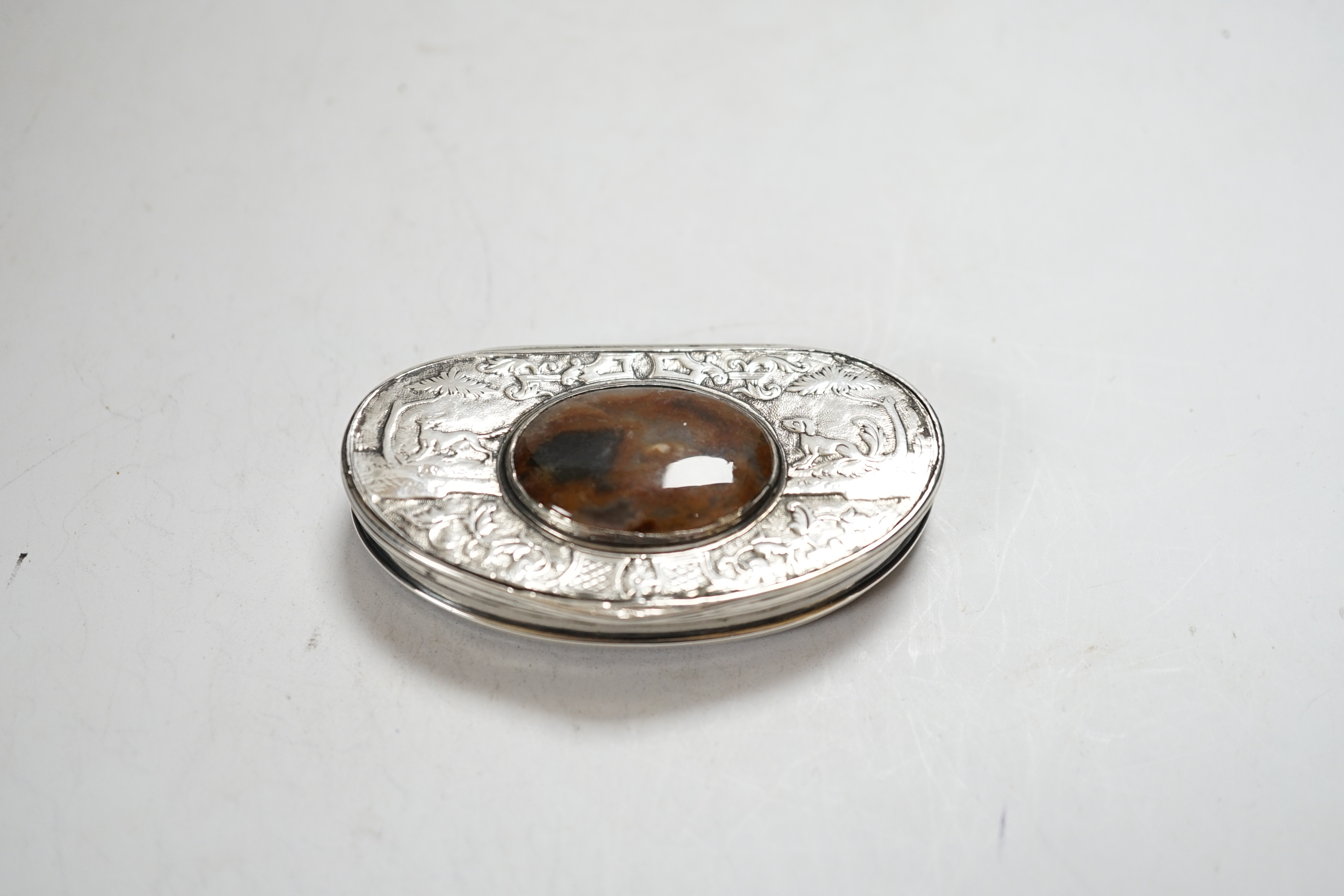 An early 19th century Scottish? white metal oval snuff box, with banded agate base and inset agate lid, decorated with dogs and scrolls, 69mm.